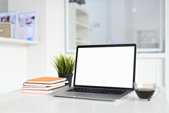 Mockup image of workspace with blank screen laptop and coffee cup on white table with blurred office room background.