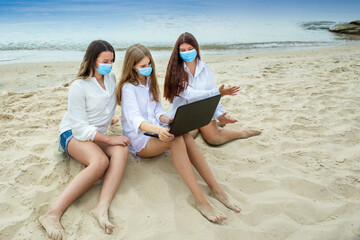 Rules for wearing a fabric medical face mask when communicating due to the coronavirus COVID 19. Girls in fabric masks
video chatting while sitting on the beach. Vacation with friends at sea.