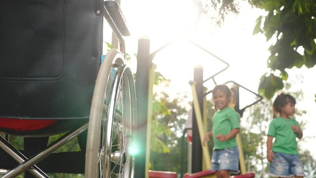 Happy disabled woman sitting in a wheelchair and watching her daughter play on exercise equipment in the outdoor public park.