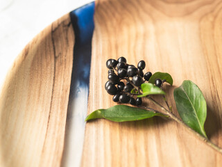 Round wooden craft tray with a blue resin insert and a branch with black berries and green leaves on white textile background. Copy space