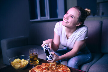 Young woman playing game at night. Nice positive cheerful model playing game with gamepad. Junk food and beer on table. Having fun alone. Dark room.