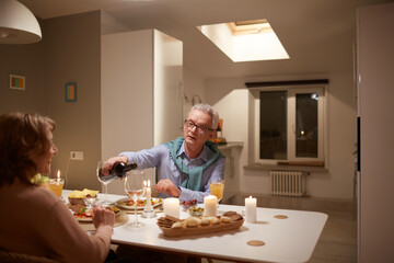 Senior man pouring the red wine into the glasses while having dinner with his wife at the table in the room