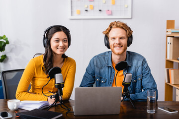 young radio hosts in wireless headphones looking at camera near microphones in studio