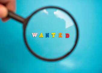 Search for employees to work. The word WANTED in colored wooden letters on a blue background through a magnifying glass.