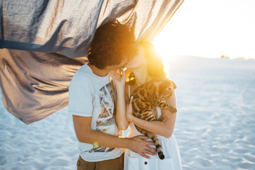 A handsome man with curly hair gently hugs his beloved. They have a cat in their hands