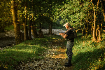 Fisherman is getting ready to start fishing in a mountain river. He is wearing special clothes and he has fishing equipment.