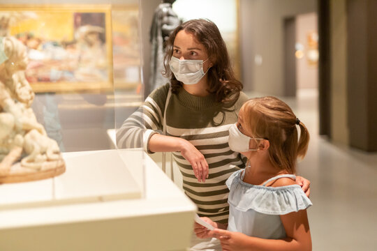 Happy cheerful mother and daughter in protective medical masks looking at expositions in museum
