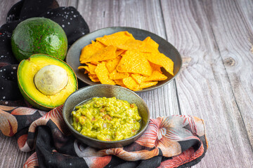 Guacamole with corn tortilla chips in plate and avocados in basket on wooden table