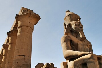 Colossus of Pharaoh Ramses II sitting in front to tall columns on throne carved with ancient...