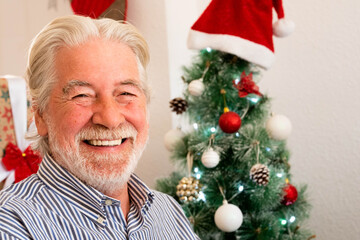Obraz na płótnie Canvas portrait and close up of old mature man smiling and laughing looking at the camera with a christmas tree at the background - old pensioner people at christmas day at home