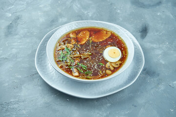 Asian ramen soup with boiled egg, noodles, mushrooms and chicken in white bowl on gray background