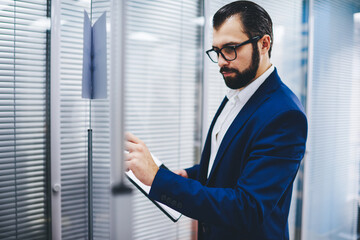 Confident bearded proud ceo in glasses making notes for financial reports, concentrated executive director dressed in formal wear writing benchmark information of working process standing in office