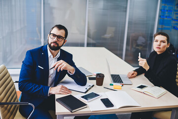 Pensive male and female colleagues in formal clothing sitting at table desktop and thinking about...