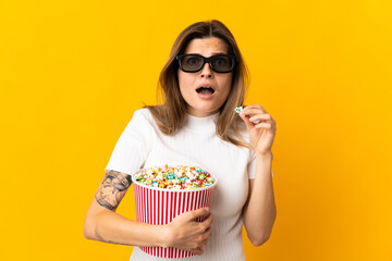 Young slovak woman isolated on yellow background with 3d glasses and holding a big bucket of popcorns