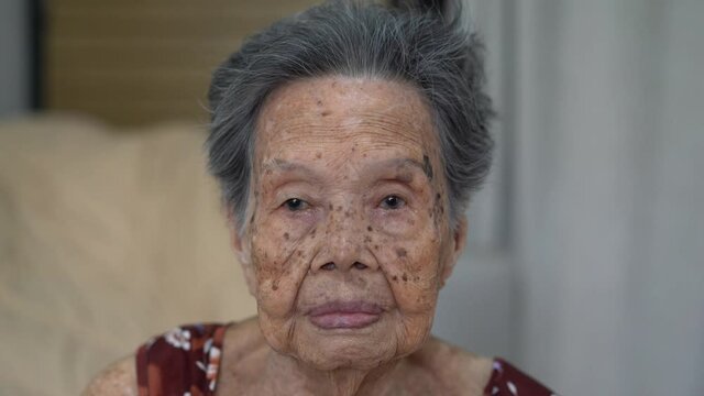 Close up portrait of elderly Asian woman smiles and waves at the camera in living room at home. Old age, gesture and people concept
