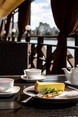 At the cafe by the lake. Close-up on a black table is a tea set with fruit tea, a black plate with cheesecake, mint leaf and cutlery.