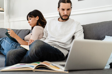 Image of serious beautiful couple reading book and working with laptop