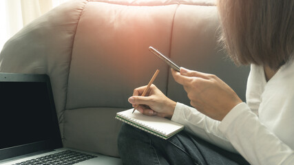 Women hands holding a smartphone and writing on a book sitting on the grey sofa in front of open laptop while working from home in quarantine for Covid-19 outbreak. Work from home concept.