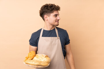 Male baker holding a table with several breads isolated on beige background looking to the side