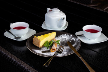Close-up on a black table is a tea set with fruit tea, a black plate with cheesecake, mint and cutlery.