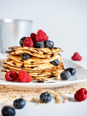 Stack of pancakes with forest fruit, raspberry, blueberry, chocolate scirope and icing sugar on a light-colored plate. White background.