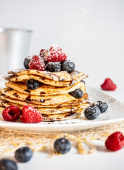 Stack of pancakes with forest fruit, raspberry, blueberry, chocolate scirope and icing sugar on a light-colored plate. White background.