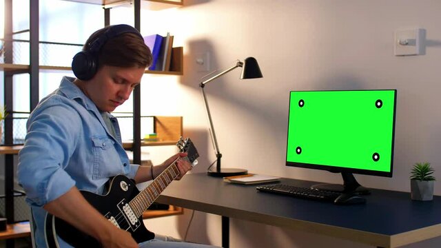 leisure, music and people concept - young man or musician in headphones with green chroma key screen on computer playing bass guitar sitting at table at home