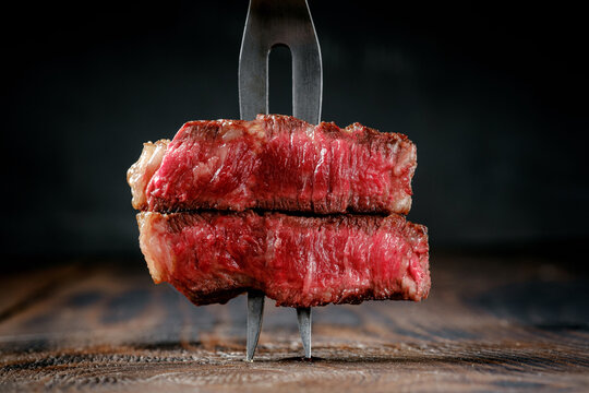 Fork with pieces of grilled striploin beef steak stuck in a wooden table, rare roasting close up