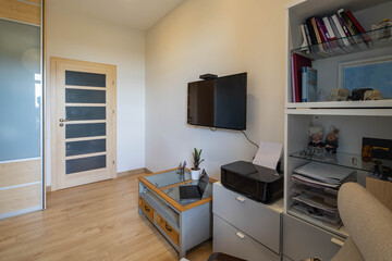 Contemporary interior of living room in modern apartment. Laptop and printer. Sliding door wardrobe. TV on wall. Wooden door.