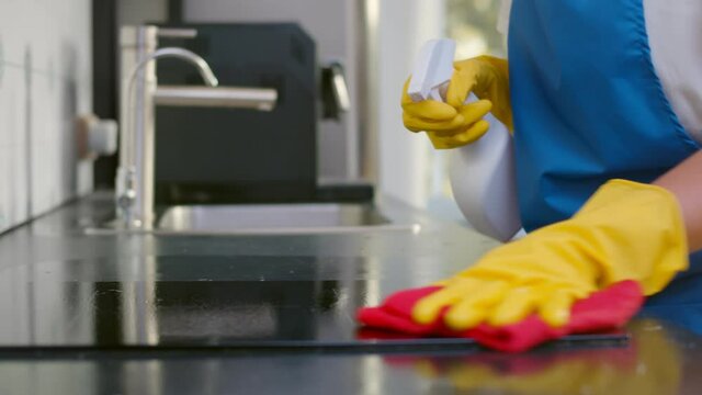 Close up of woman in gloves and apron spraying detergent and using clothe cleaning oven panel