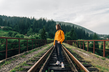 Portrait of fashionable woman tourist in casual clothes standing on railway bridge against mountains background with forest and posing for camera. Hipster travels. Vorokhta, Ukraine.