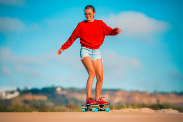 Sporty happy carefree woman in blue shorts, red sneakers and hoodie riding longboard in sunny summer day. Holidays fun.