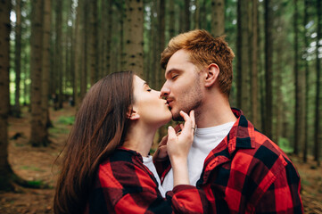 Close-up photo. Young man and woman in red shirts kissing in the woods with closed eyes. A loving couple kisses and hugs on a walk in the woods.