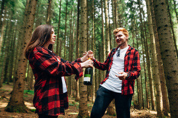 Happy couple in red shirts opening champagne in the woods on a walk. Woman tears off a bottle of wine in the woods on a walk with her boyfriend.