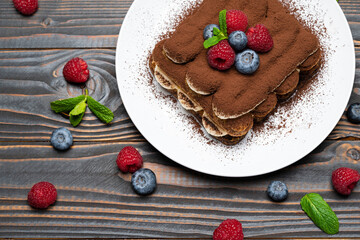 portion of Classic tiramisu dessert with raspberries and blueberries on wooden background