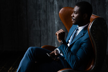 African American businessman in a blue classic suit sits in an aviator loft armchair with a phone in his hands