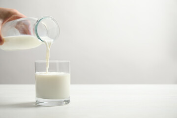 Woman pouring milk into glass at white table, closeup. Space for text