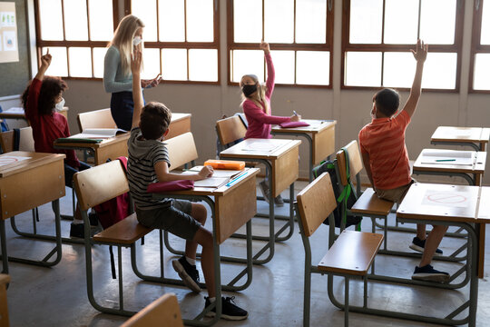 Group Of Kids Wearing Face Masks Raising Their Hands In The Class