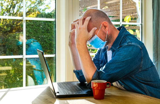 Stressed out man sitting by his laptop with his hands in his face while wearing a face mask.
