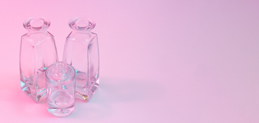 Concept with two shaped square-bottom and one cylindrical round-bottom crystal clear clean empty glasss bottles standing on colorful background in blue and pink neon vibrant lights. Copy space.