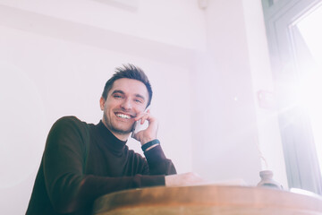 Positive young man speaking on smartphone while sitting at table near window in cafe