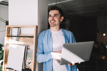 Cheerful Caucasian male copywriter smiling while waiting for laptop updating in coworking space,...