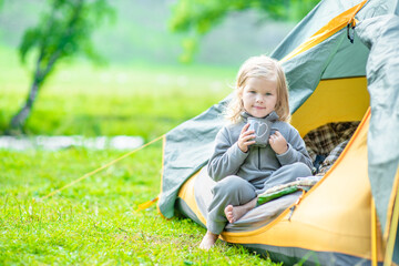 Little girl sits inside a tent at morning and holds cup of a tea. Empty space for text