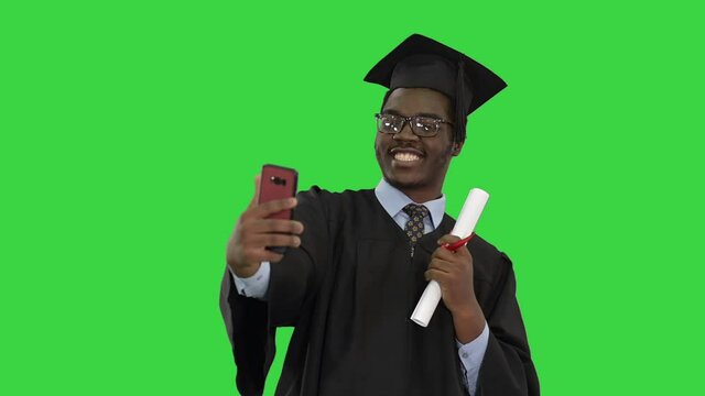 Happy african american male student in graduation robe taking phone selfies with his diploma on a Green Screen, Chroma Key.