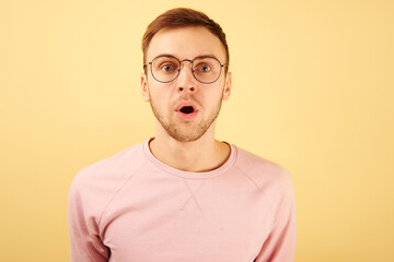 Portrait of shocked guy surprised with open mouth, emotional caucasian face with glasses in pink casual clothes on yellow background isolated