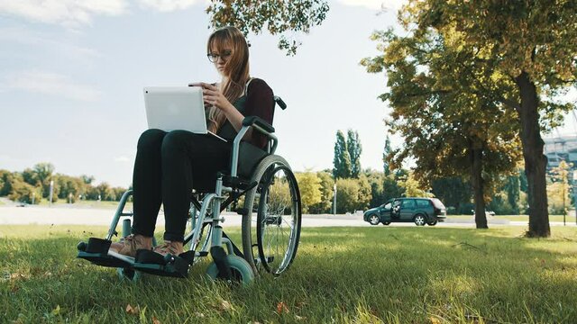 Remote work concept. Young disabled woman in the wheelchair using smartphone and laptop. High quality 4k footage
