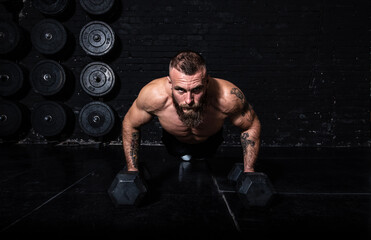 Fototapeta na wymiar Young sweaty strong fit muscular man with big muscles doing push ups on the dumbbells in the gym as hardcore cross workout training with free copy space