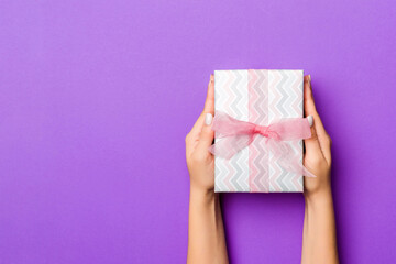 Flat lay of woman hands holding gift wrapped and decorated with bow on purple background with copy space. Christmas and holiday concept