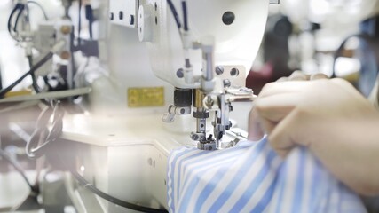 Sewing on machine, hands and blue and white striped cloth close up. Sewing process, female hands on white machine, blue shirt on table with white sewing machine