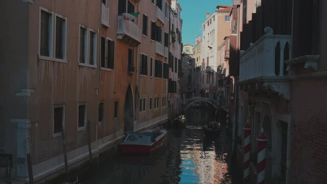 European trip. Dark narrow canal street with parked gandolas by the buildings. People crossing the bridge in distance.